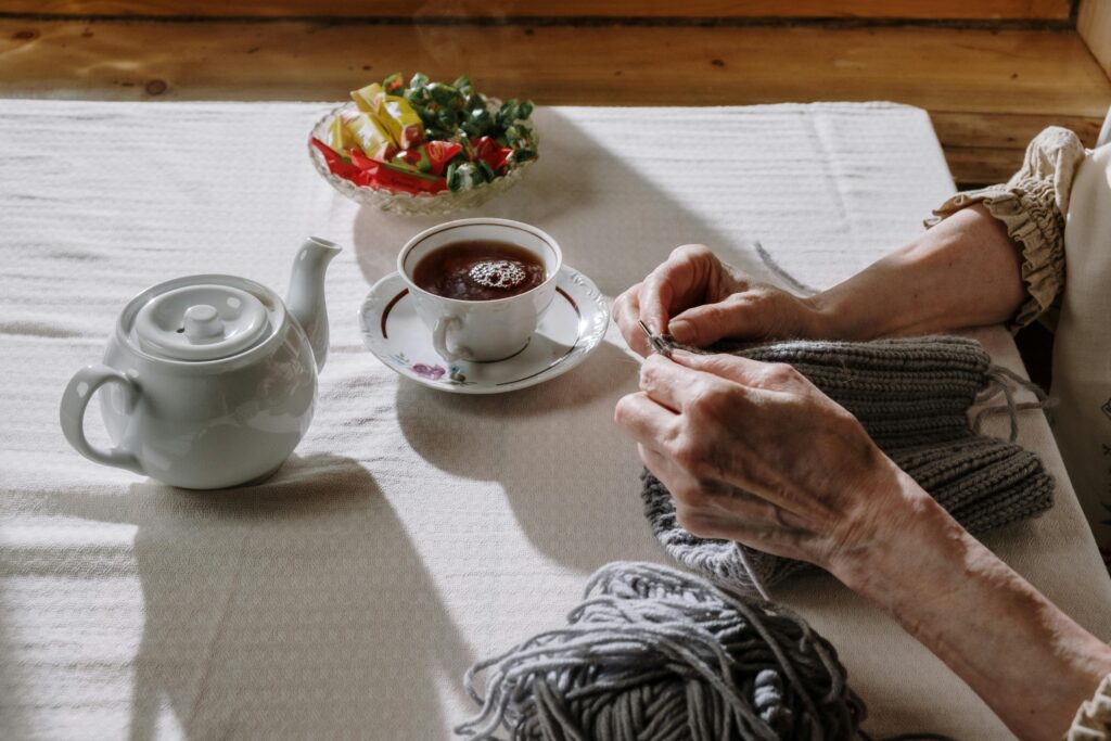 A senior woman knitting at a table with a cup of tea and a teapot nearby.